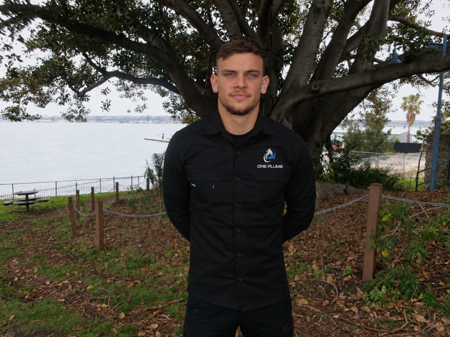 Luka from One Plumb standing under a big tree by the water in his black One Plumb shirt