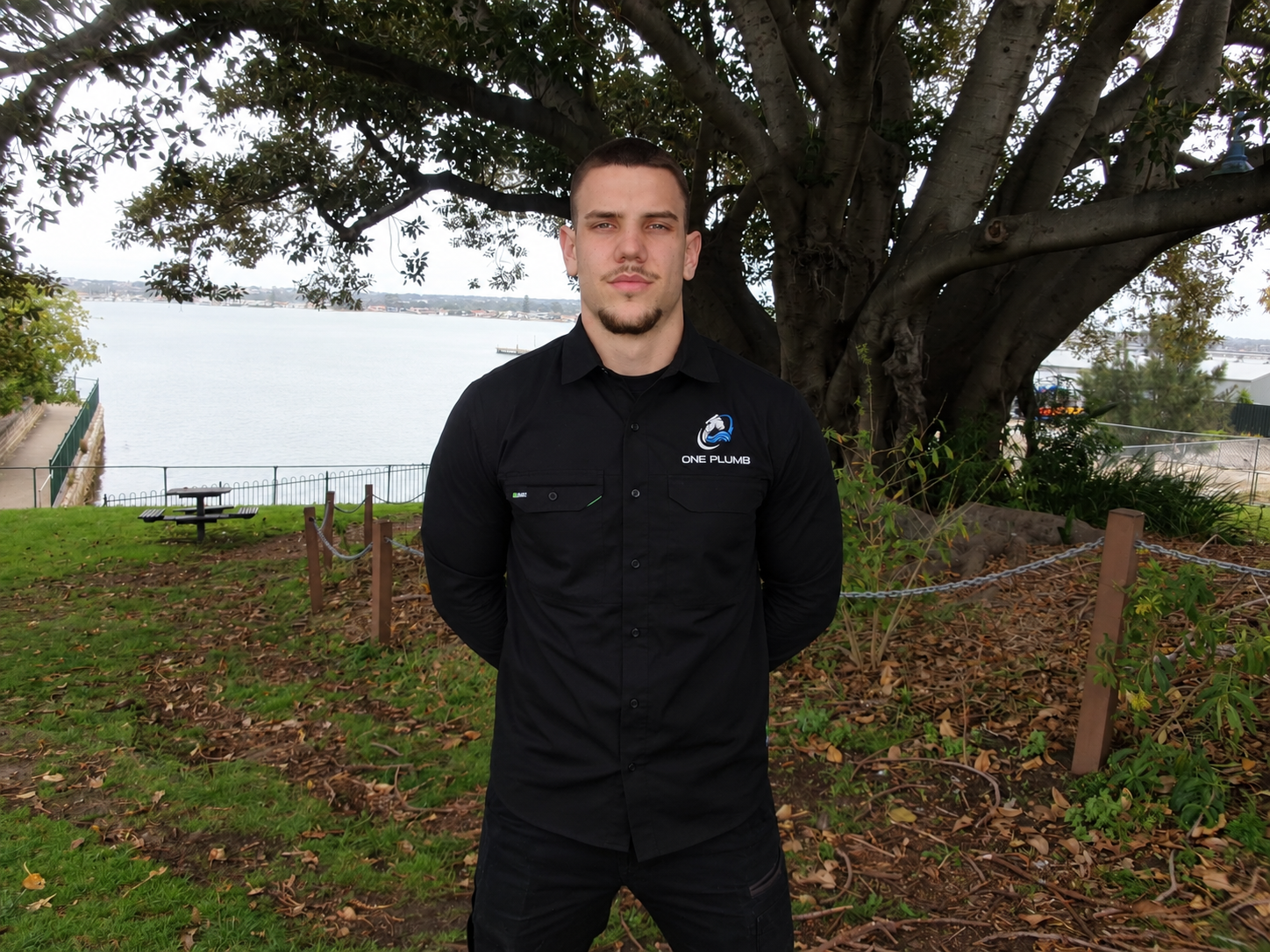 Tomi from One Plumb standing under a big tree by the water in his One Plumb shirt